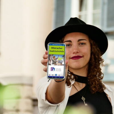 Eine junge Frau hält ein Smartphone in die Kamera. Darauf ist der digitale Stadtrundgang abgebildet.