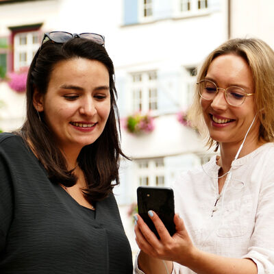 Visitors look at a smartphone. Both are smiling. One of the people has headphones.
