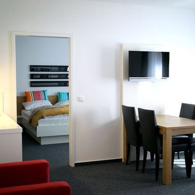Blick in moderne und helle Wohnung mit Sitzgelegenheit, Esstisch und separatem Zimmer mit Doppelbett.