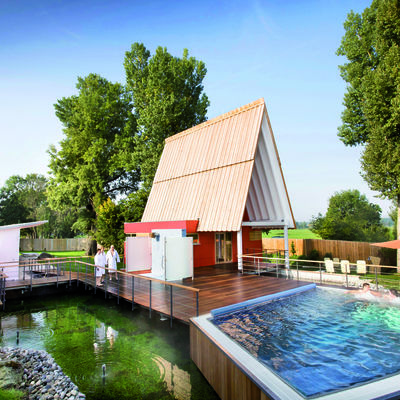 Wellnesslandschaft unter freiem Himmel. Rotes Saunahaus mit spitzem Dach. Terrasse besteht aus Holz mit integriertem Poolbecken.