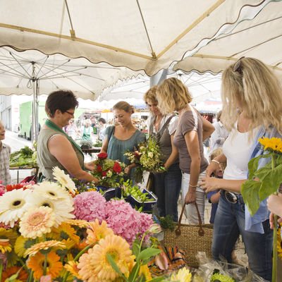 Many different and colorful cut flowers can be seen in the foreground. In the background you can see a sales situation in which a saleswoman is presenting women with a colorful bouquet.