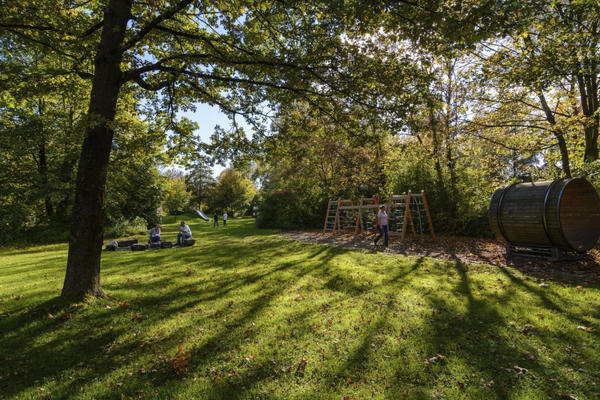 Spacious and shady meadow, framed by trees and bushes. A climbing frame for children and a barbecue area can also be seen.