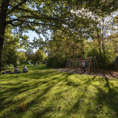 Spacious and shady meadow, framed by trees and bushes. A climbing frame for children and a barbecue area can also be seen.