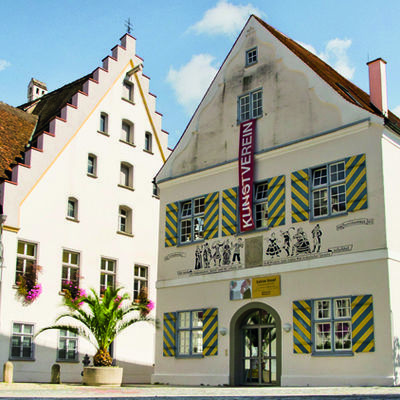 Large house without roof overhang. The shutters are painted obliquely black and yellow. The door is rounded at the top. Various human caricatures can be seen above the door in the middle as an allusion to the Dramatic Society and the play by Shakespear "The Tempest", which was performed there for the first time in German.