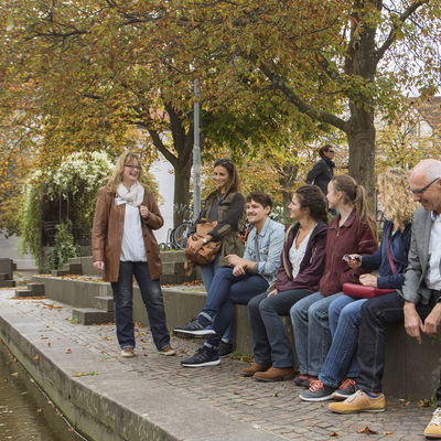 Zwei Männer und vier Frauen sitzen auf einer Mauer an einem Stadtbach und hören einer Stadtführerin zu, die vor ihnen steht.