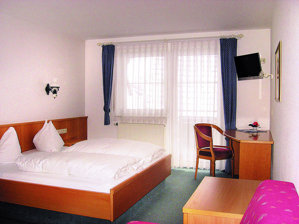 Hotelzimmer mit Doppelbett, Schreibtisch und Schrank sowie großem Fenster mit Gardinen.