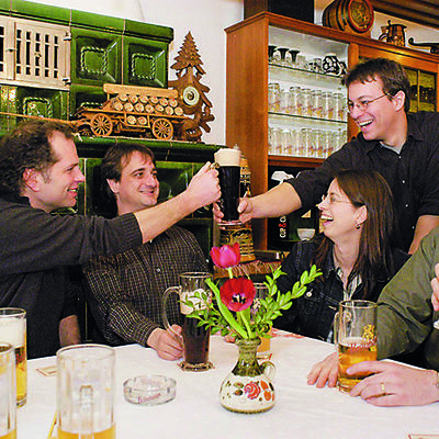 Um einen Tisch in einer Wirtschaft sitzen mehrer Personen die fröhlich dreinblicken, vor ihnen steht Bier. Zwei Männer prosten sich zu.