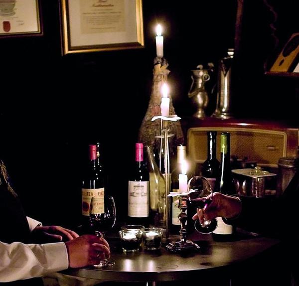 Ein Tisch im dunkeln, beleuchtet mit drei Kerzen. Weihnflaschen sowie ein gefülltes Weinglas stehen auf dem Tisch. Das Weinglas wird von einer Person berührt. 