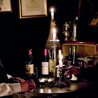 Ein Tisch im dunkeln, beleuchtet mit drei Kerzen. Weihnflaschen sowie ein gefülltes Weinglas stehen auf dem Tisch. Das Weinglas wird von einer Person berührt.