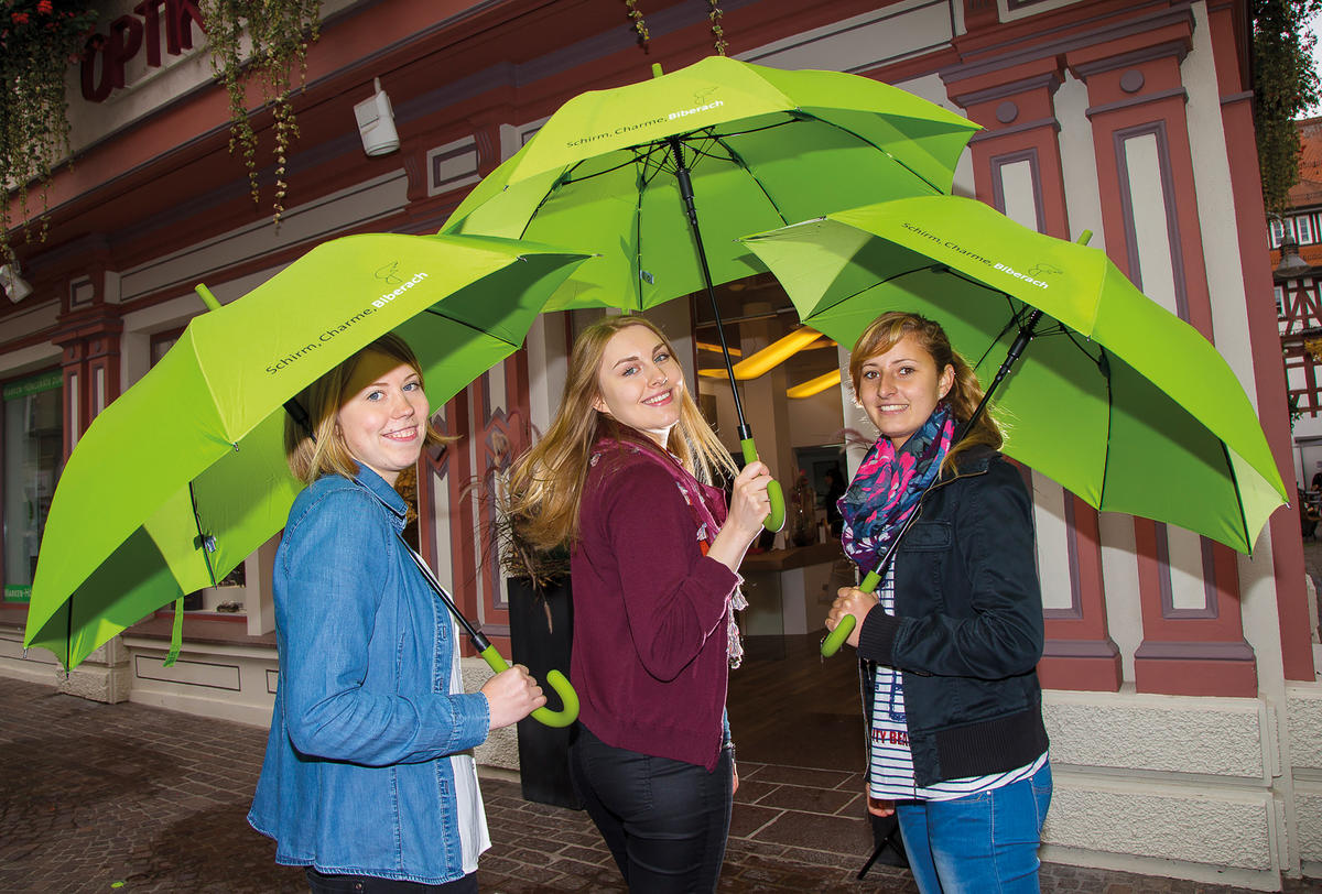 Drei junge Frauen mit grünem Schirm in der Hand schauen lächelnd in die Kamera. Den Leihschirm gibt es für 10 Euro an der Tourist-Info im Rathaus.