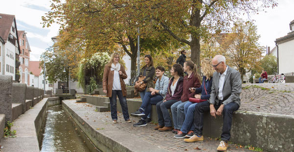 Das Bild zeigt Teilnehmer einer Stadtführung. Es ist eine Gruppe von sieben Personen. Sechs davon sitzen auf einer erhöhten Stufe aus Stein an einem Stadtbach. Die Stadtführerin steht vor der Gruppe und spricht mit begeistertem Gesichtsausdruck. 