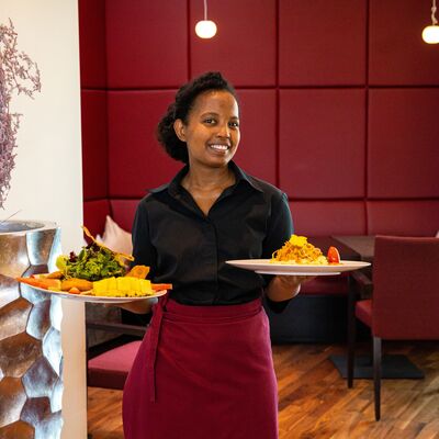 Restaurant Ropach. Eine Mitarbeiterin des Restaurant Ropach hält zwei Teller mit Speisen in der Hand und lächelt freundlich aufs Kamera.