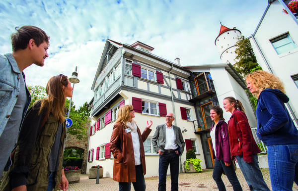 A city tour takes place in front of a renovated half-timbered house with red shutters. The group is in a semicircle around the city guide.